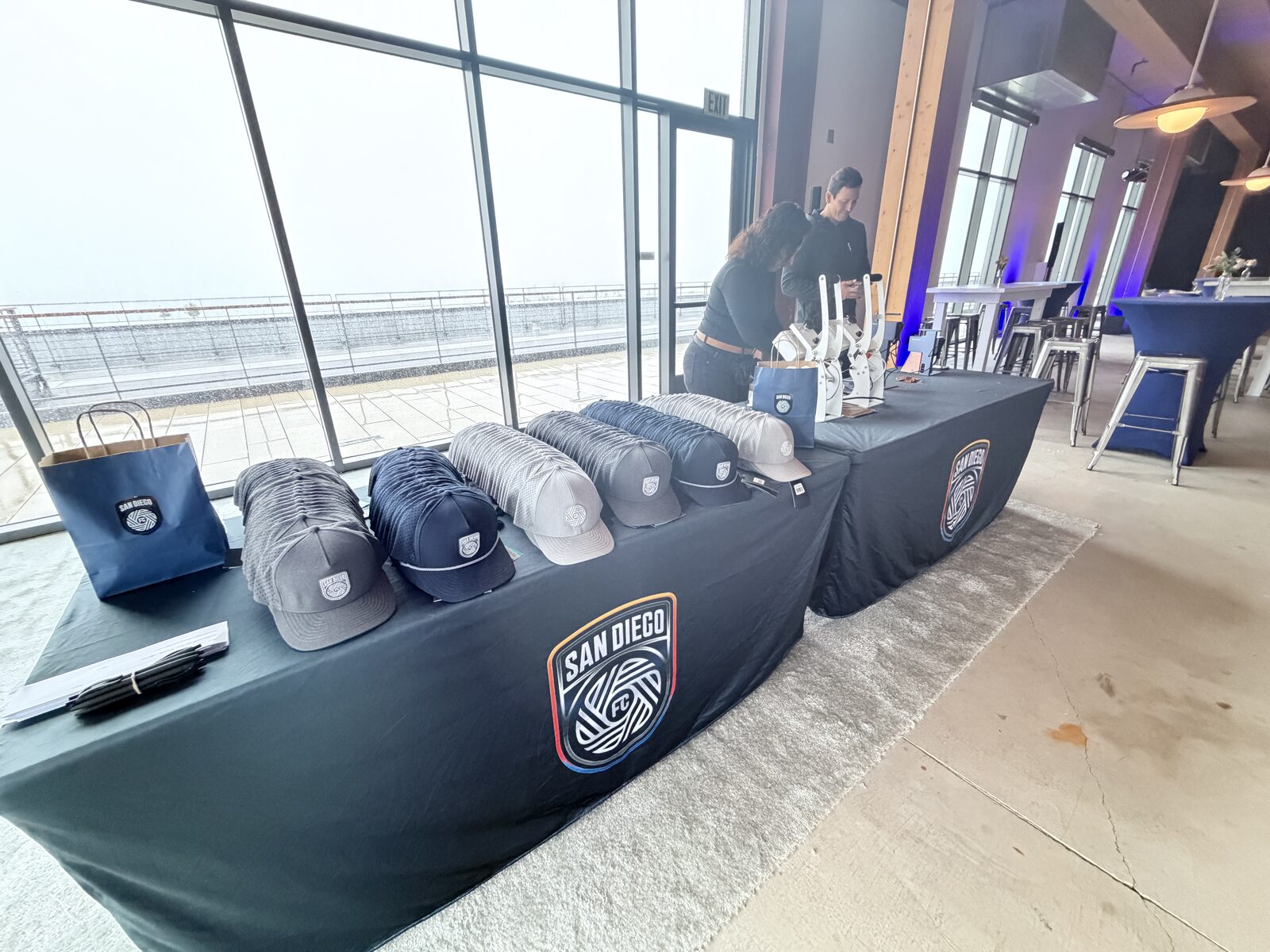 Rows of gray, navy, and cream trucker caps lined up on a branded table before doors at a soccer club lounge